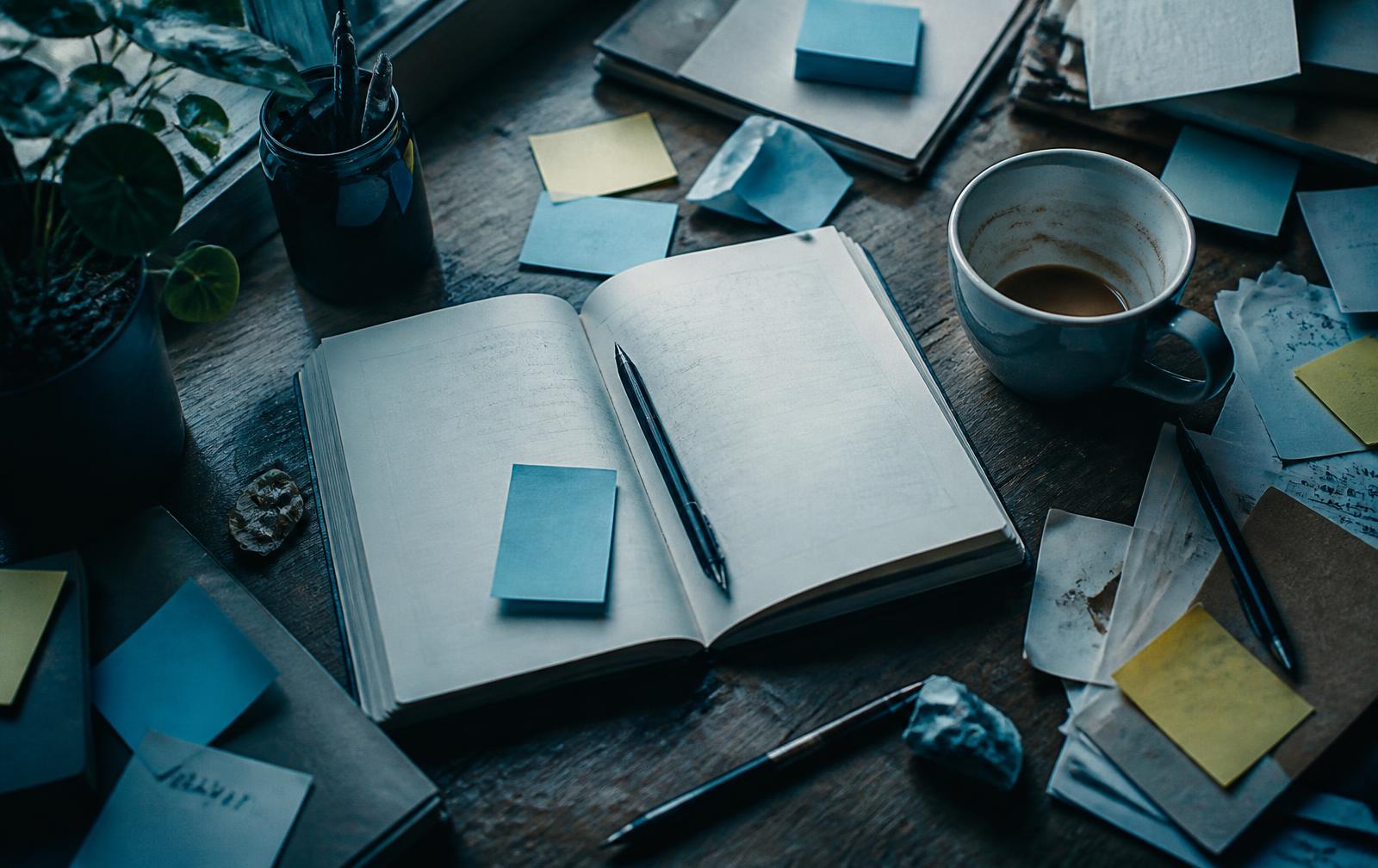 A cluttered desk with scattered notes, an open notebook, and cold coffee